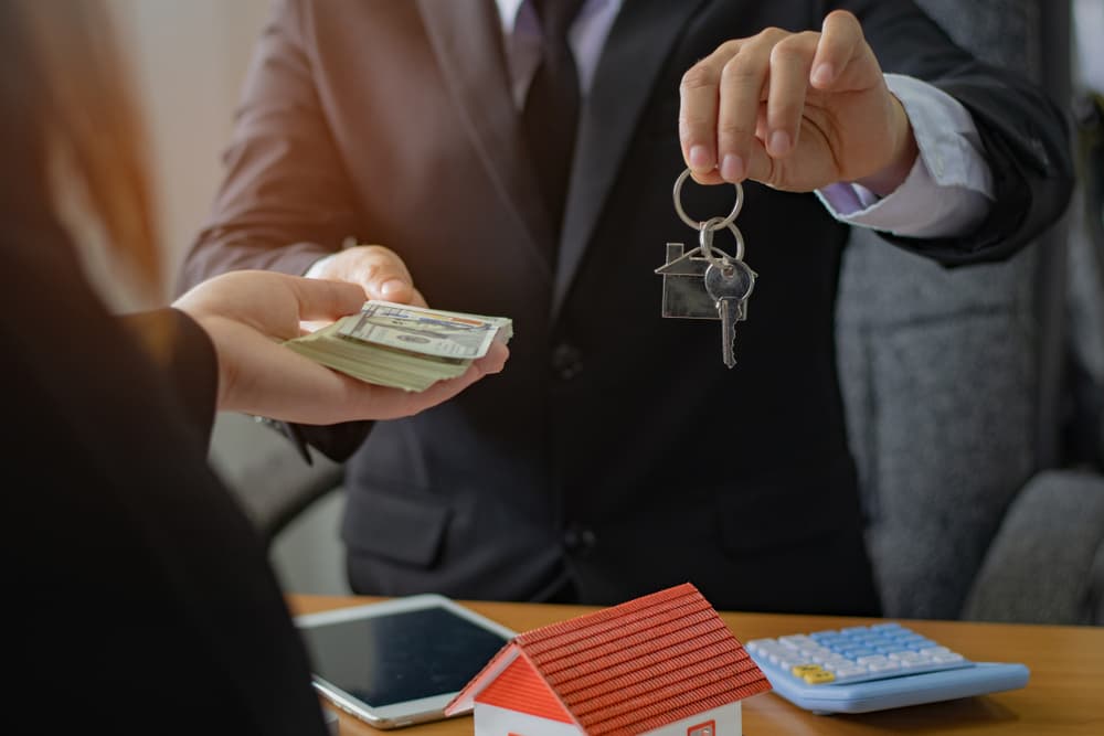 Real estate agents exchanging keys and holding a US dollar banknote at agency office, symbolizing agreement concept.