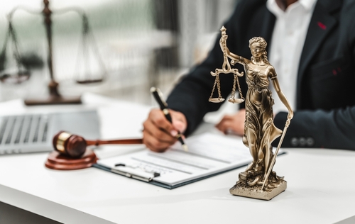 Attorney signing official legal documents beside a bronze Lady Justice statue and a gavel on a bright office desk.