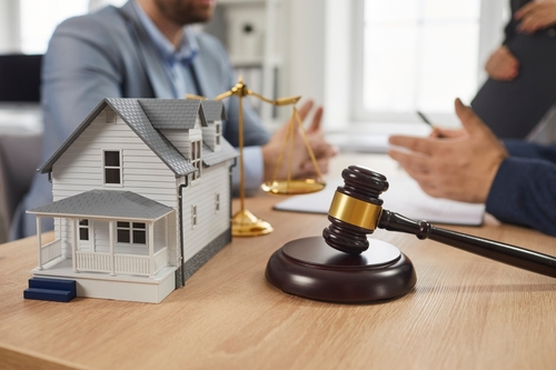 Close-up of a miniature house model and a judge’s gavel on a desk while two people discuss legal documents in the background, representing property law or real estate legal consultation.
