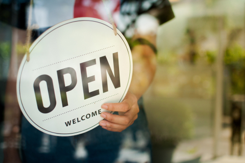 Person holding a round “OPEN” sign on a glass door, welcoming customers into a small business storefront.