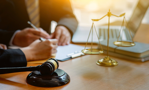 lose-up of a judge’s gavel and brass scales of justice on a wooden desk while two people review legal documents and take notes in the background.