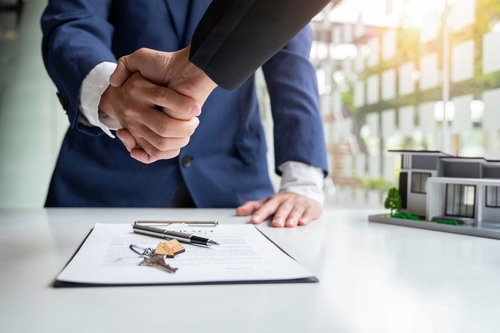 Two people in business attire shaking hands across a desk with real estate documents, keys, and a small house model.