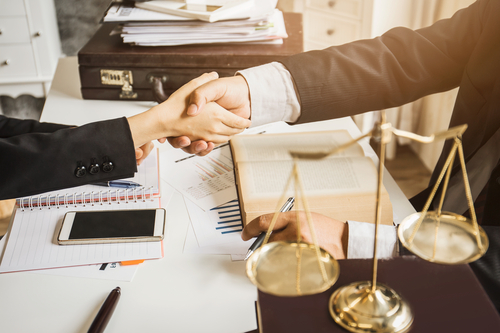 Close-up of two people shaking hands across a lawyer’s desk with legal documents, scales of justice, and a gavel in view.