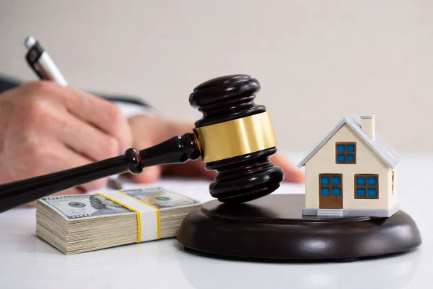 A house and gavel sit on a desk next to some cash, while a person signs a contract or other document in the background.